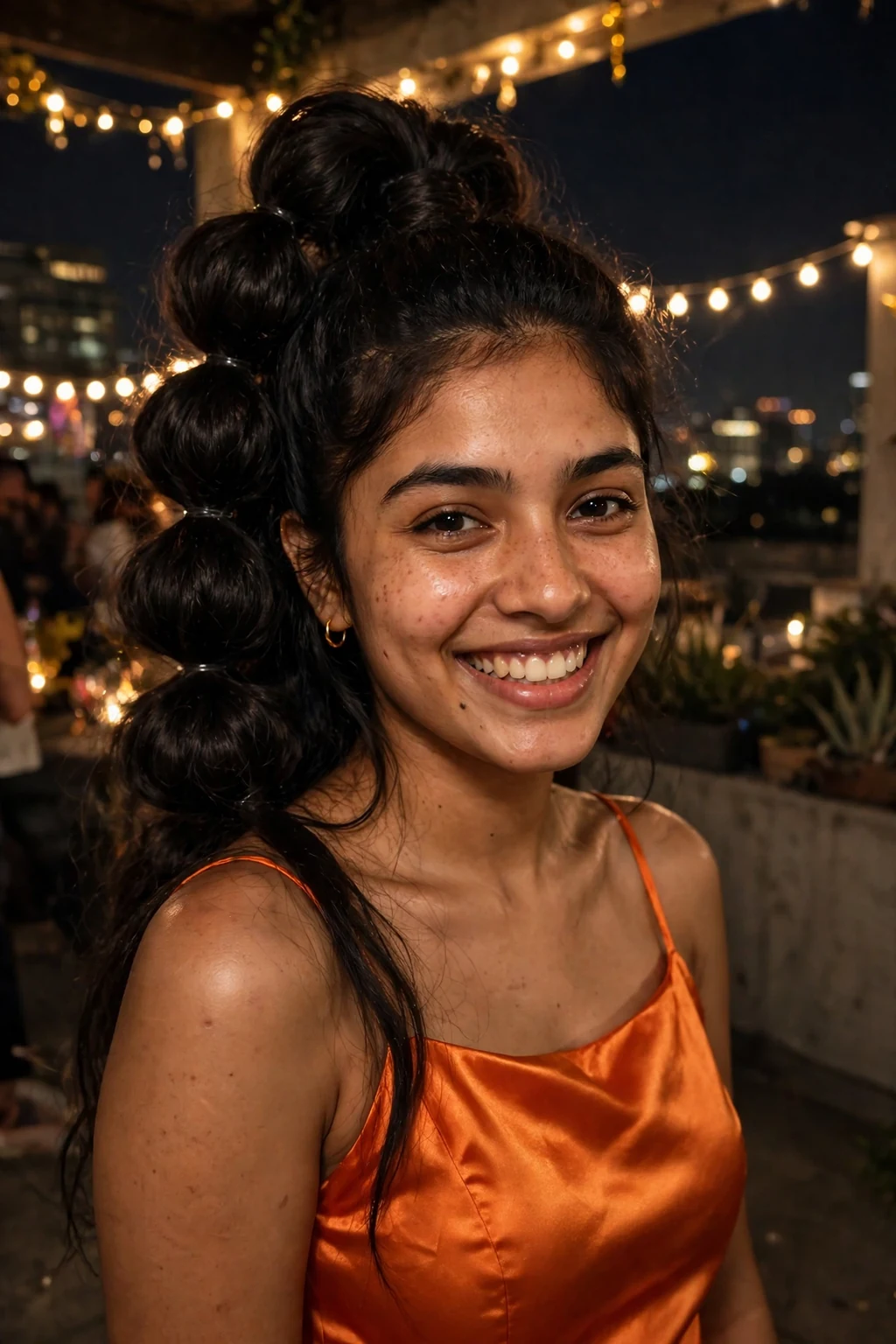Young South Asian woman with a bubble ponytail wearing an orange satin dress at a rooftop party.