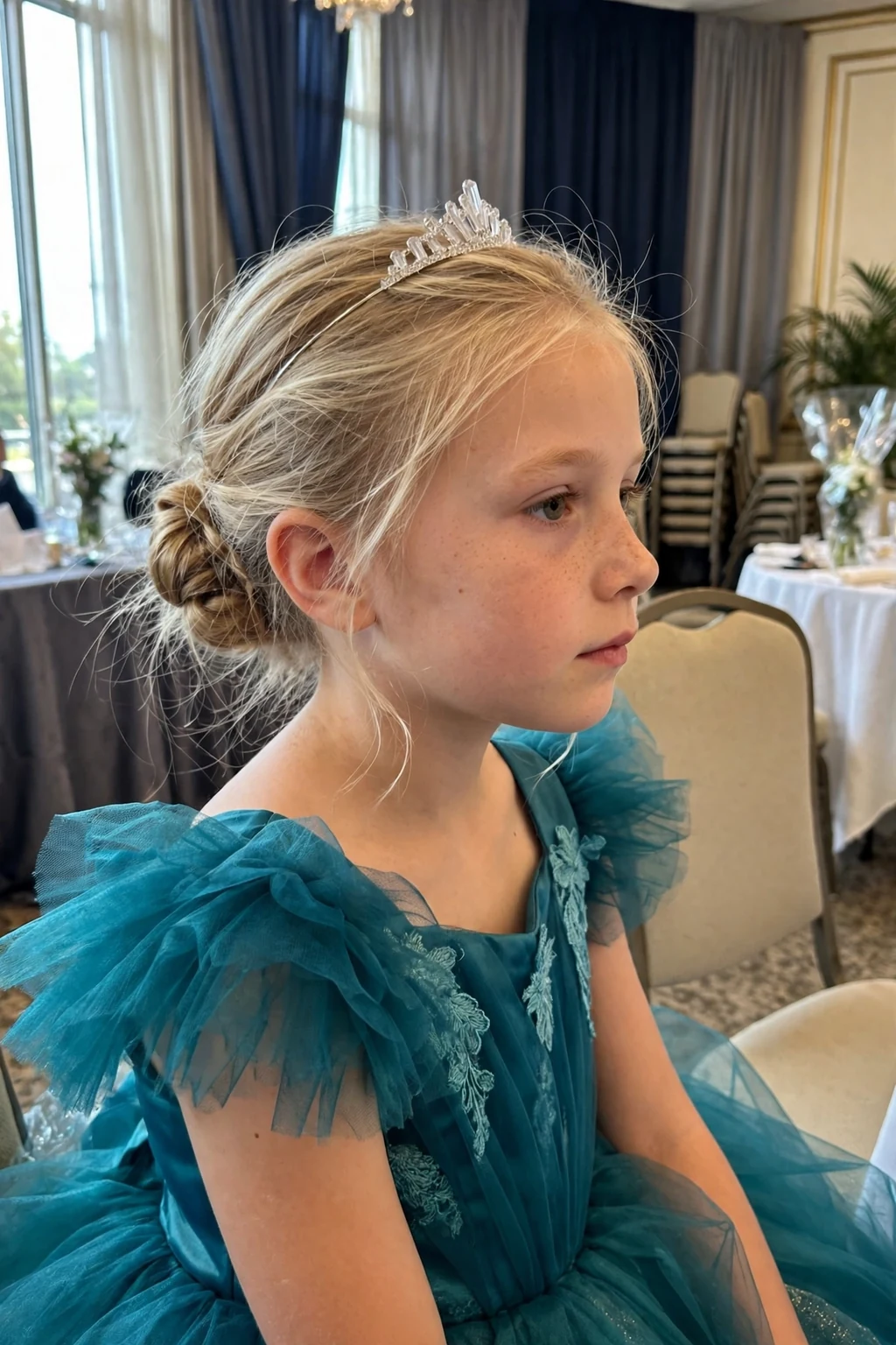 A young girl with a low chignon and a crystal tiara in a hotel ballroom before a party.