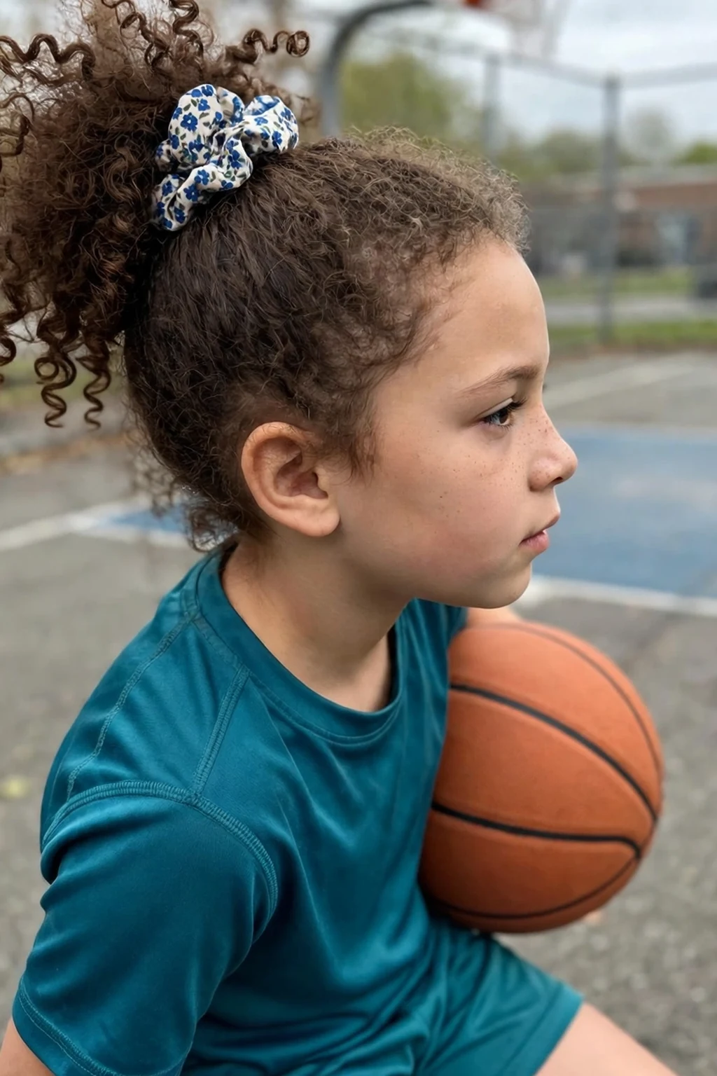 A young girl with a high curly puff held by a patterned scrunchie, on a basketball court.