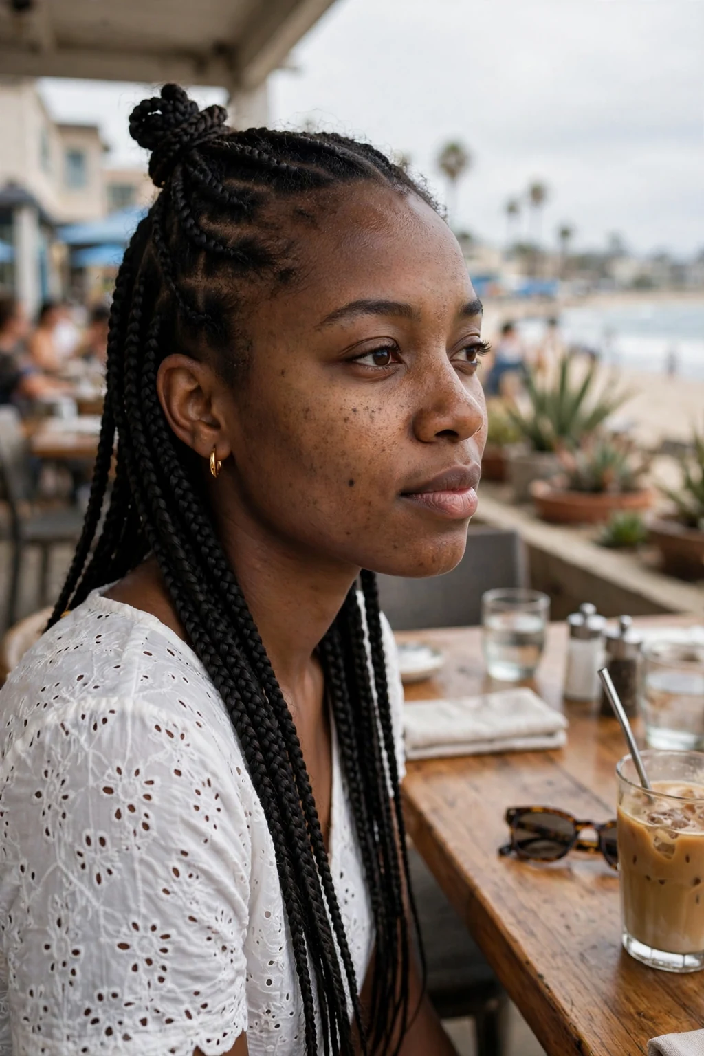 Young Black woman with knotless box braids in a half-up top knot with the rest falling loose, photographed in a beachside cafe wearing a white eyelet top.