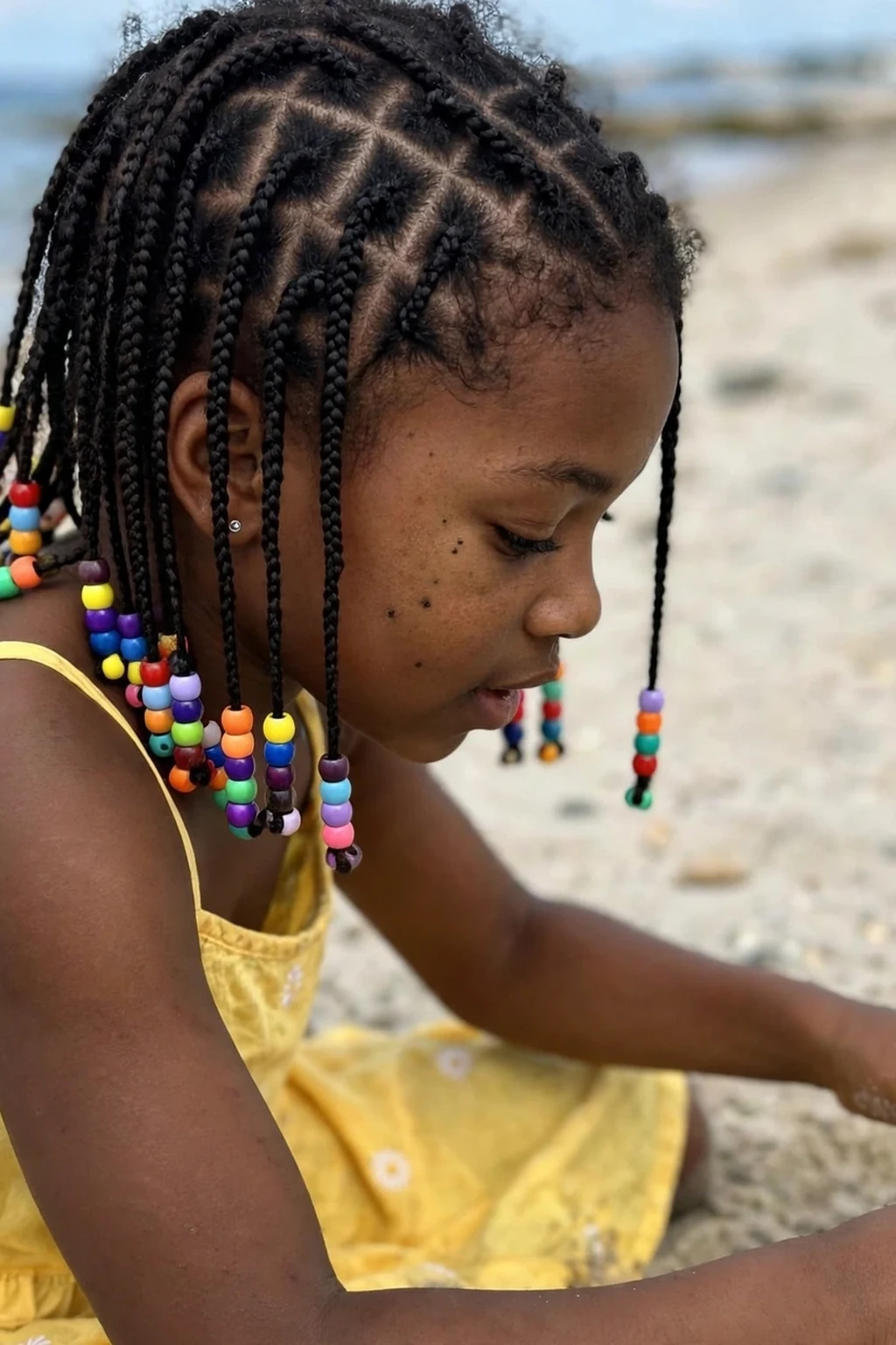 A young girl with many mini braids finished with colorful beads, playing on a sandy beach.