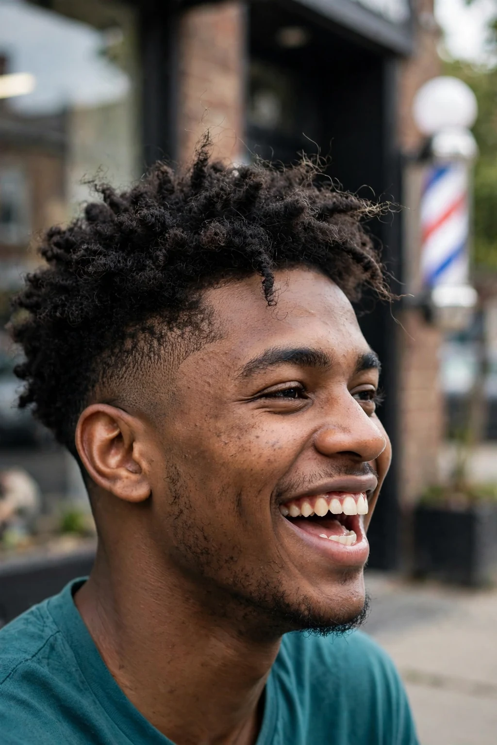 Young Black man with short coils and a fade for thick hair, laughing outside a barbershop in a teal shirt.