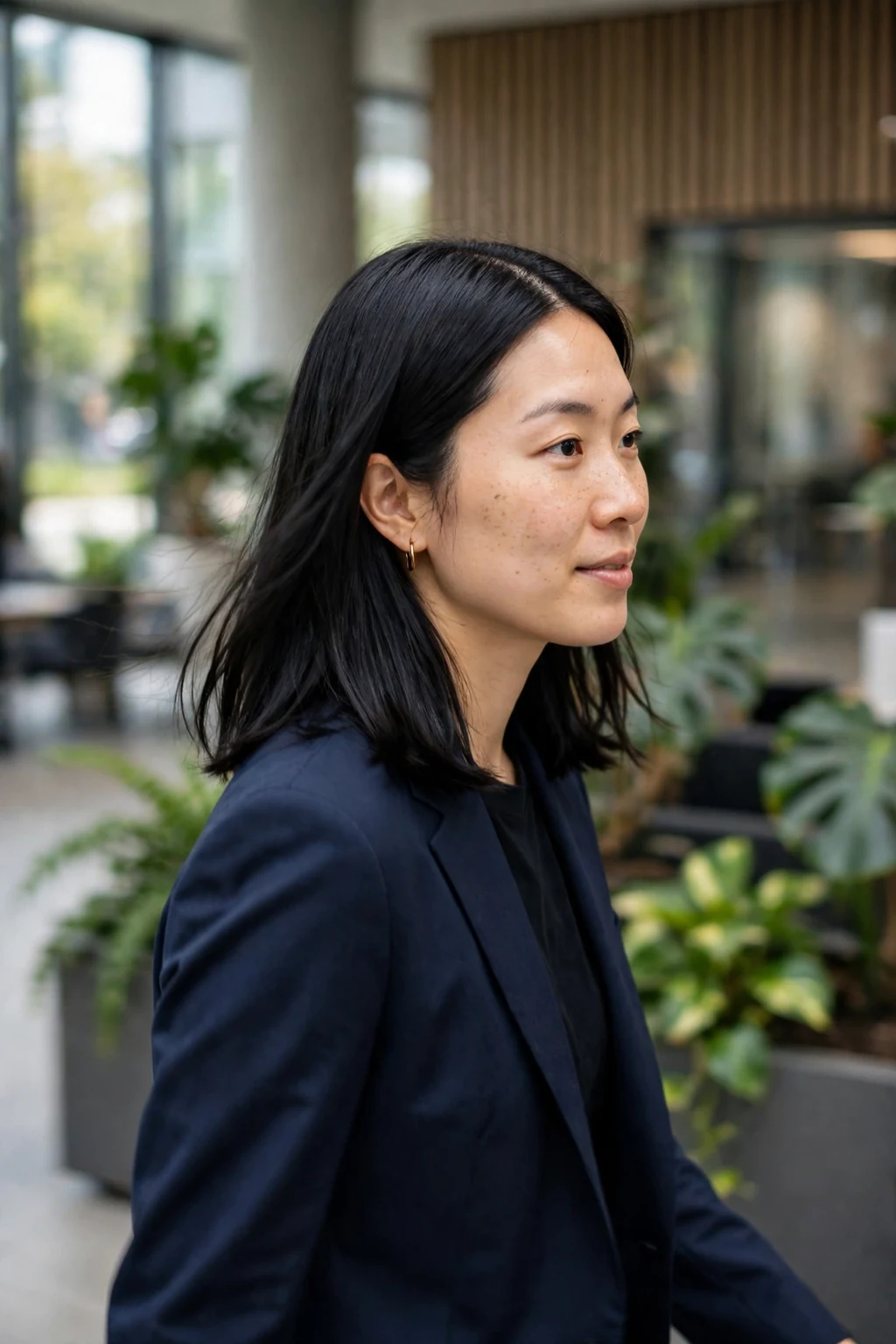 A young woman with a sleek middle-part lob and glass hair shine, in a modern office lobby wearing a navy suit.