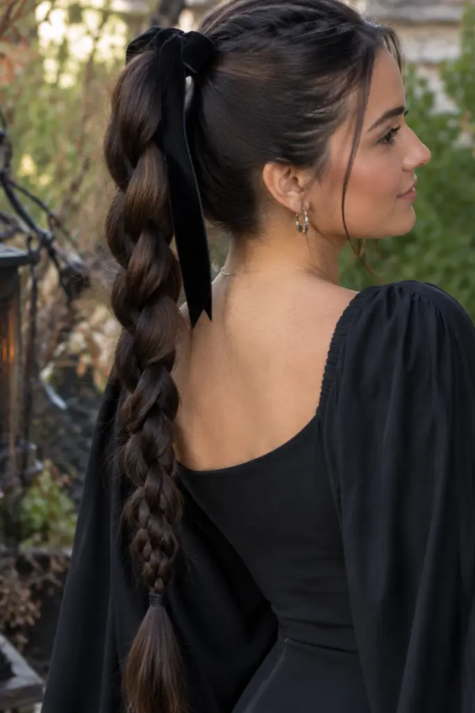 young woman with long dark brown hair in a high ponytail and thick loose braid tied with a black velvet ribbon, wearing a flowing black dress in a warm Halloween garden.